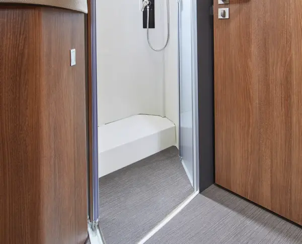 Ein modernes Badezimmer mit einer teilweise geöffneten Holztür, die einen weißen Duschbereich mit einer Glasabtrennung, einer Handbrause und einem grauen Bodenbelag zeigt, der in den Duschbereich führt.