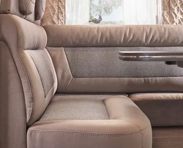Eine Nahaufnahme einer modernen, gepolsterten Wohnmobil-Sitzgruppe mit braunfarbener Sitzfläche, Rückenlehne und eingebautem Tisch. Das Sonnenlicht fällt sanft durch ein Fenster und leichte Vorhänge im Hintergrund.