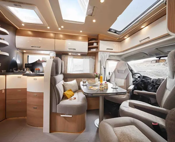 Modernes Wohnmobil-Interieur mit grauen und hölzernen Oberflächen, einem Esstisch für das Frühstück, bequemen Sitzgelegenheiten, großen Fenstern, Oberlichtern und einem Blick durch die Windschutzscheibe auf die Wüstenlandschaft.