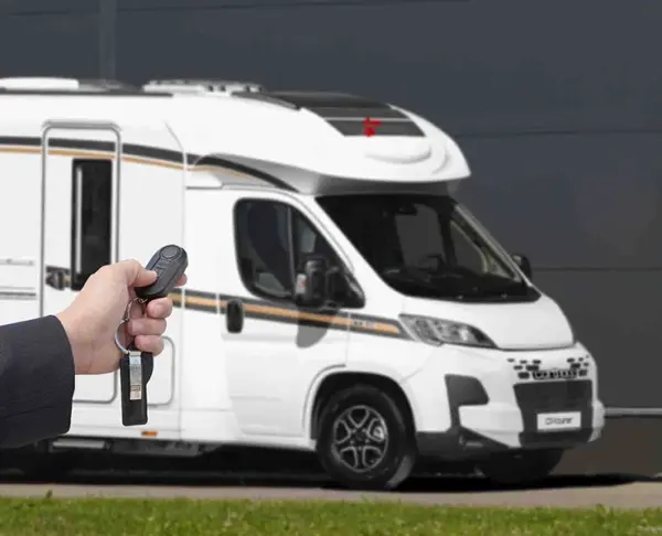 Eine Person hält einen Schlüsselanhänger mit Fernbedienung in der Hand, der auf ein weißes Wohnmobil gerichtet ist, das in der Nähe auf einer gepflasterten Einfahrt mit einer dunklen Wand im Hintergrund geparkt ist.