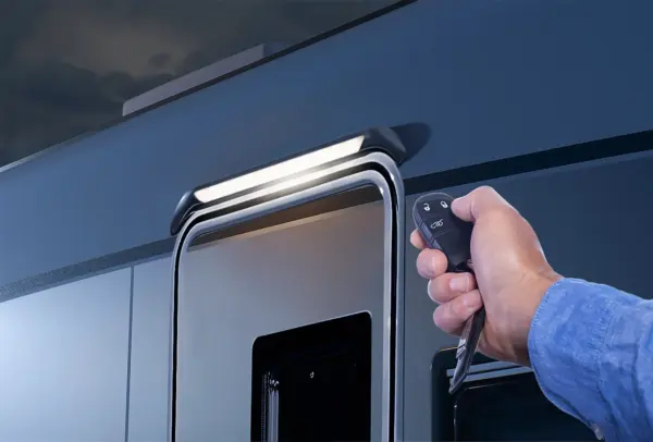 Eine Person hält einen Schlüsselanhänger mit Fernbedienung in die Nähe der Tür eines modernen Wohnmobils Carthago Chic S Plus, über der in der Dämmerung eine Außenleuchte leuchtet.