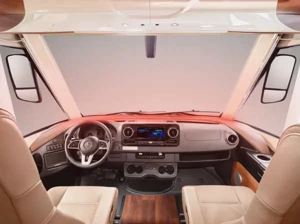Blick ins Innere eines Wohnmobils mit zwei beigen Kapitänssesseln, einem modernen Armaturenbrett mit Touchscreen-Display, einem Lenkrad mit Mercedes-Benz Logo, Becherhaltern und Holzakzenten.