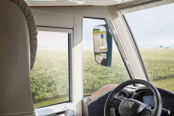 Blick aus dem Inneren eines Fiat-Fahrzeugs mit Lenkrad, Seitenfenster und Spiegel, in dem sich ein ländliches Straßenschild spiegelt, während draußen ein Feld und der Himmel zu sehen sind.