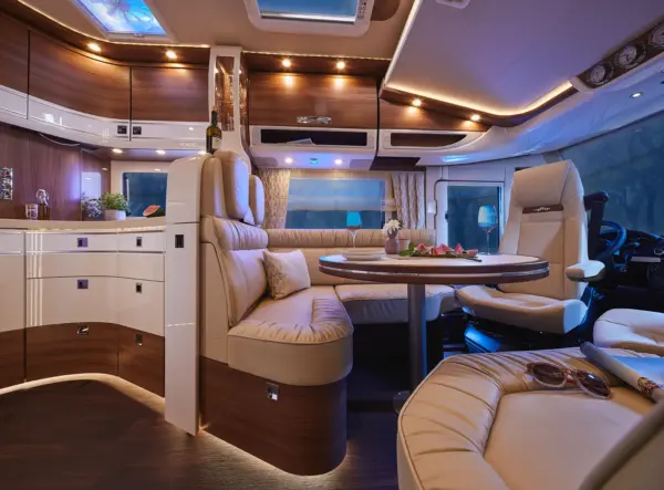 Luxuriöses Wohnmobil-Interieur mit braunen Ledersitzen, rundem Esstisch mit Tellern und Blumen, gewölbten Schränken, stimmungsvoller Beleuchtung und großen Fenstern mit Vorhängen.