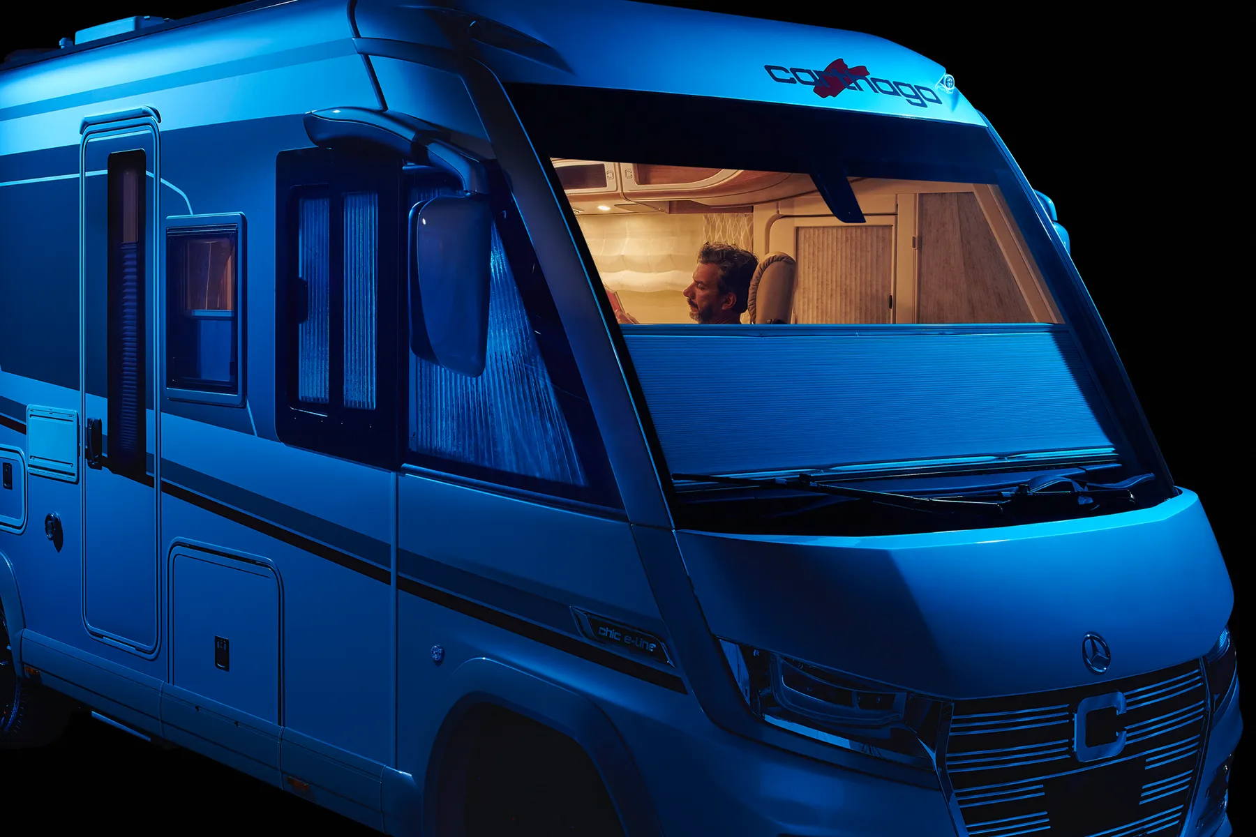Ein Mann sitzt nachts in einem modernen Wohnmobil, das von warmen Innenleuchten beleuchtet wird. Die vordere Windschutzscheibe des Fahrzeugs wird teilweise von einer Jalousie verdeckt. Die Szene spielt sich vor einem schwarzen Hintergrund ab.