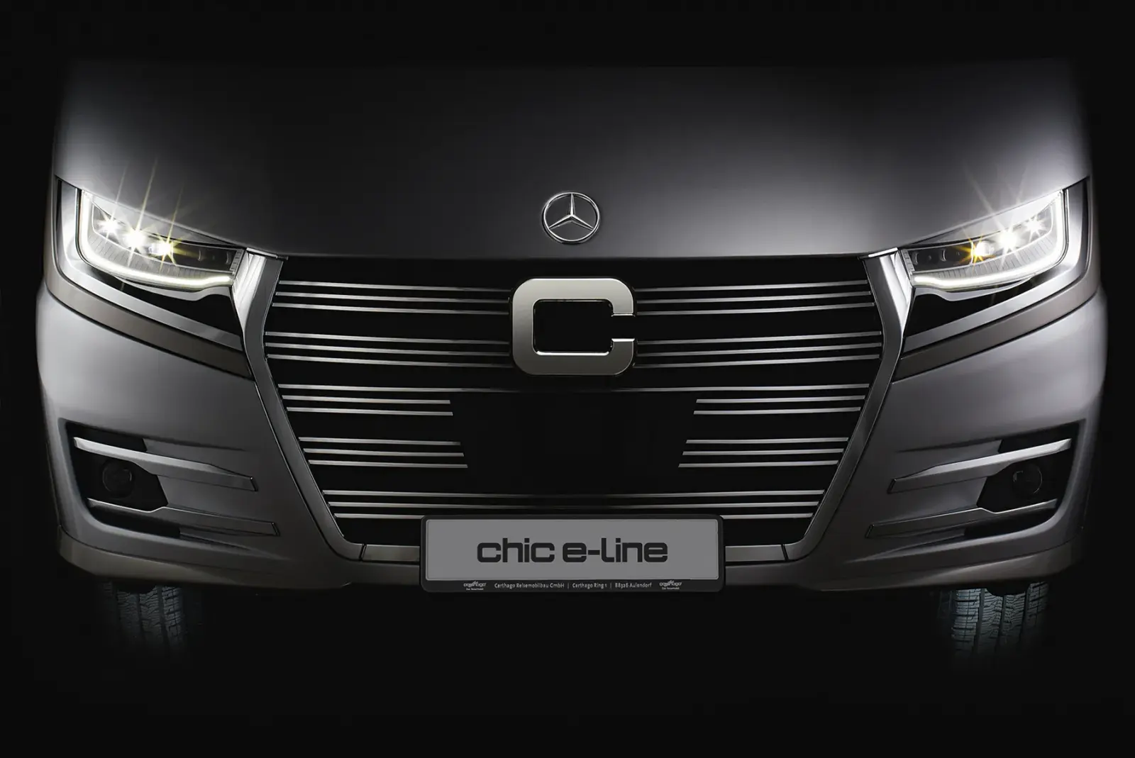 Eine Nahaufnahme des Kühlergrills und der Scheinwerfer eines eleganten, modernen Fahrzeugs mit der Bezeichnung e-line, mit einem markanten C-Emblem und beleuchteten Scheinwerfern in dunkler Umgebung.