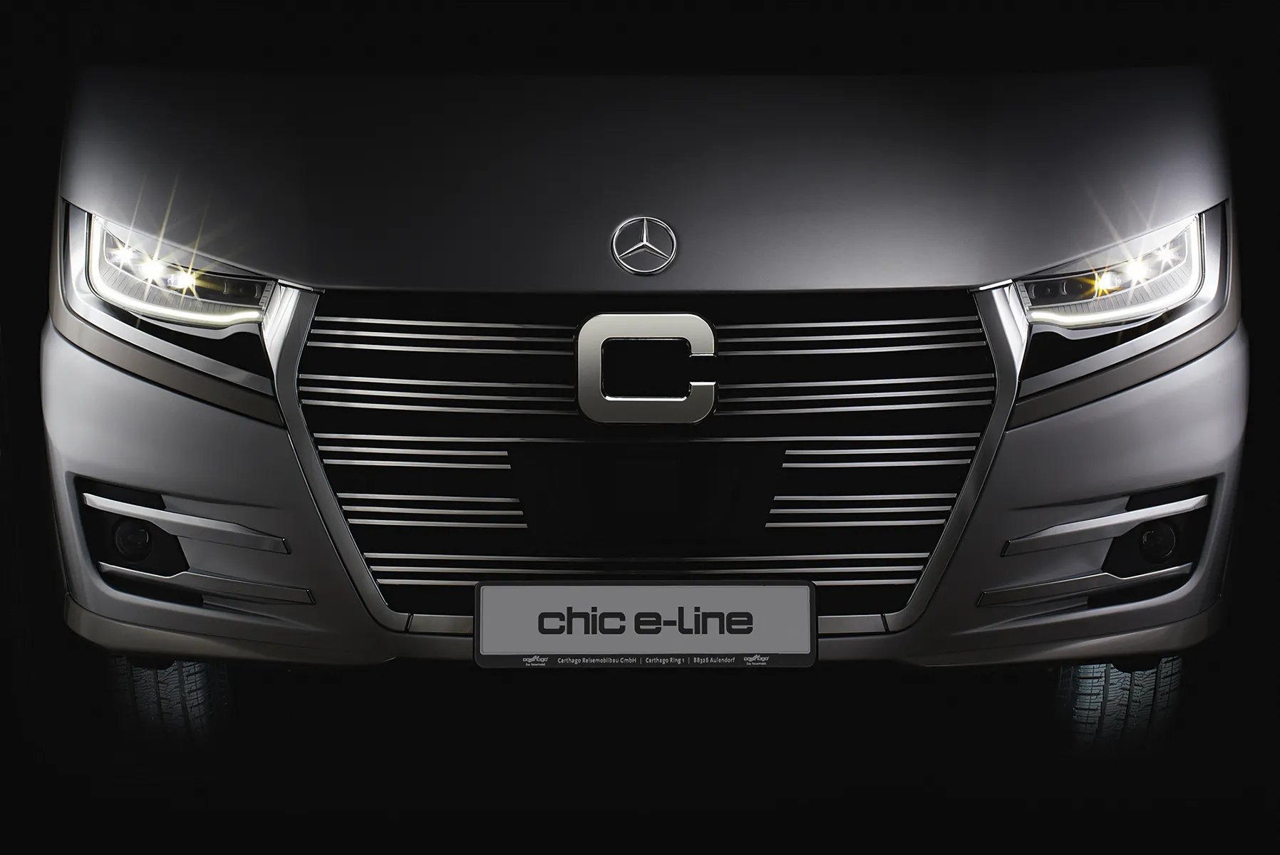 Eine Nahaufnahme des Kühlergrills und der Scheinwerfer eines eleganten, modernen Fahrzeugs mit der Bezeichnung e-line, mit einem markanten C-Emblem und beleuchteten Scheinwerfern in dunkler Umgebung.