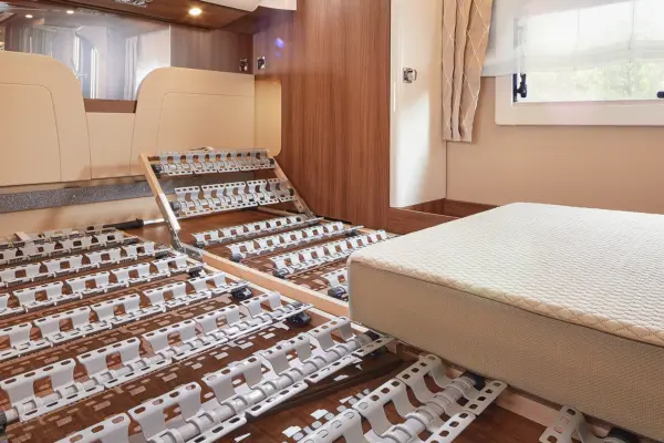 Ein modernes Wohnmobil-Interieur mit einem verstellbaren Metall-Lattenrost, bei dem ein Teil hochgeklappt ist, einer Matratze auf der rechten Seite und Holzwänden mit einem großen Fenster, das Tageslicht hereinlässt.