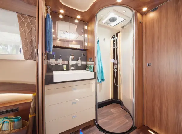 Modernes Wohnmobil-Badezimmer mit Holzvertäfelung, einem weißen Waschbecken mit einem großen Spiegel darüber, reichlich Stauraum in den Schubladen und einer Dusche mit Glasabtrennung. Blaue Handtücher hängen an der Wand, und eine sanfte Beleuchtung schafft eine gemütliche Atmosphäre.