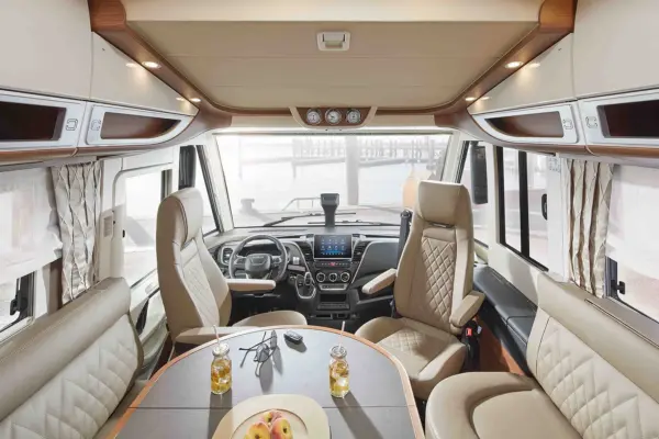 Luxuriöses Wohnmobil-Interieur mit hellbraunen Ledersitzen, einem Lenkrad, einem Armaturenbrett und einem Esstisch mit zwei Getränken und einer Obstschale, große Fenster bieten einen hellen, malerischen Blick nach draußen.