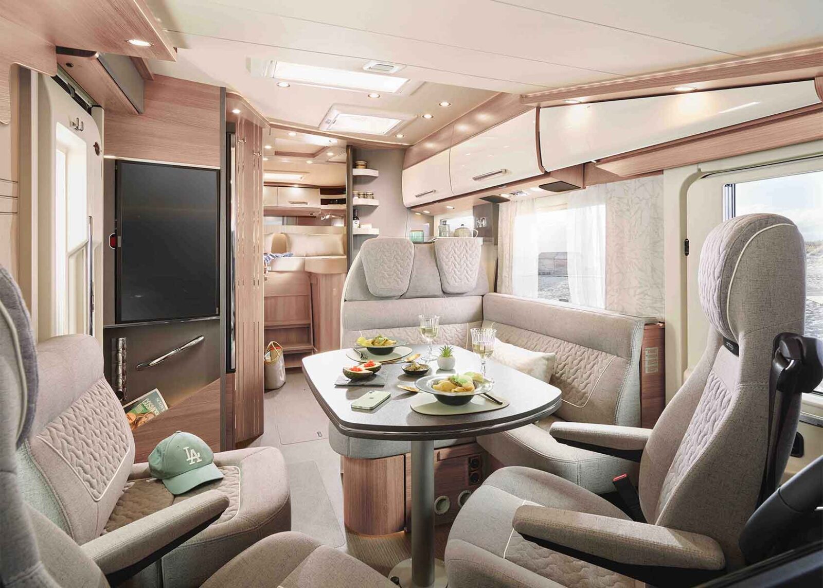 Modernes vollintegriertes Wohnmobil-Interieur mit graubraunen gepolsterten Sitzen um einen Esstisch mit Tellern mit Speisen und Getränken. Zu sehen sind Hängeschränke, eine Küchenzeile und ein Fenster, durch das Tageslicht einfällt. Auf einem Sitz befindet sich eine grüne Mütze.
