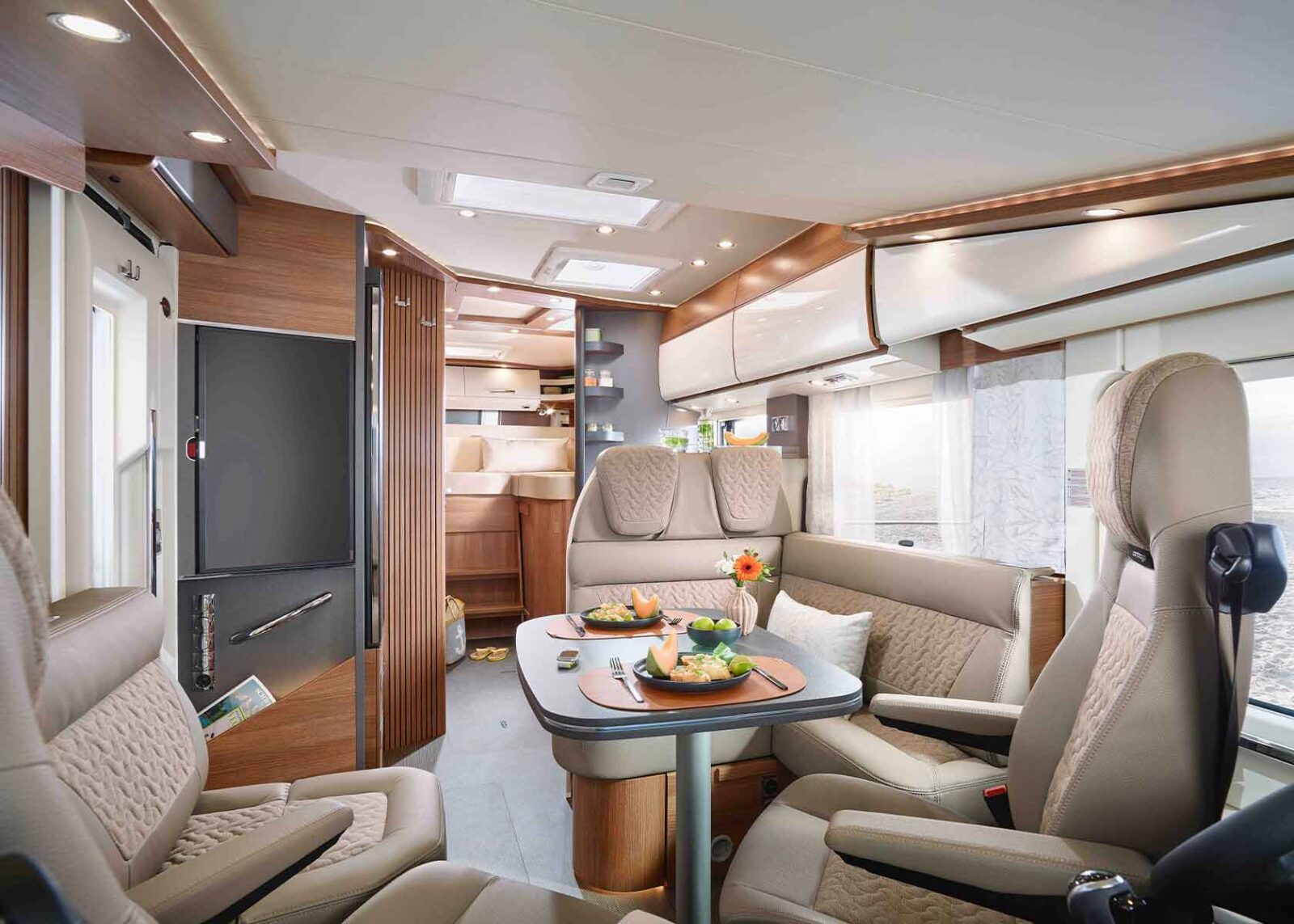 Modernes vollintegriertes Wohnmobil-Interieur mit beigen Ledersitzen, einem gedeckten Tisch mit Speisen, Blumen, Holzschränken und großen Fenstern, die natürliches Licht hereinlassen. Die Ansicht zeigt einen gemütlichen, gut beleuchteten Wohn- und Essbereich.