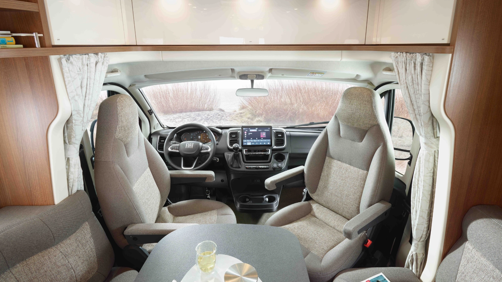Innenansicht eines teilintegrierten Wohnmobils mit Fahrer- und Beifahrersitzen, einem Lenkrad, einem Armaturenbrett mit Bildschirm, einem kleinen Tisch mit Wasser und einem Snack sowie beigen Vorhängen an den Seiten.
