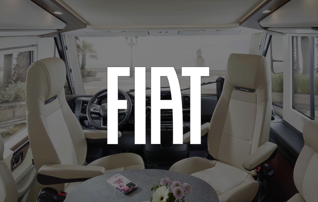 Der Innenraum eines integrierten Wohnmobils von Fiat ist mit cremefarbenen Sitzen, einem runden Tisch mit Blumen und einem großen FIAT-Logo in weißen Buchstaben ausgestattet.