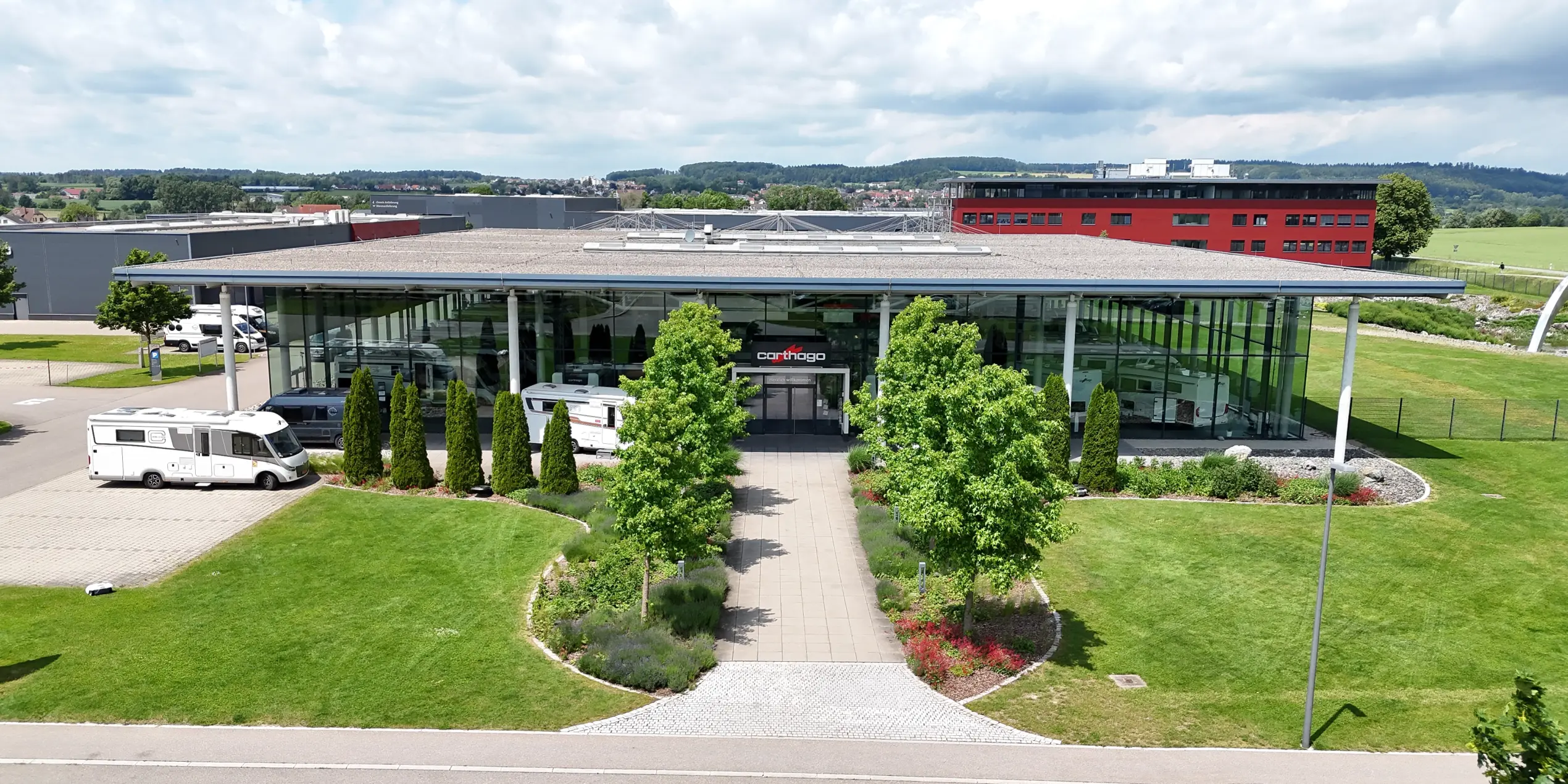 Ein modernes Glasgebäude mit Flachdach, umgeben von grünen Rasenflächen, Bäumen und ein paar geparkten integrierten Wohnmobilen. Ein gepflasterter Gehweg führt zum Eingang. Der Himmel ist teilweise bewölkt und im Hintergrund sind Industriegebäude zu sehen.