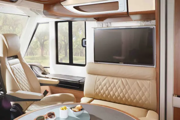 Luxuriöses Carthago Chic E Line Wohnmobil-Interieur mit beigen Ledersitzen, einem runden Tisch mit Snacks und Getränken, großen Fenstern und einem Flachbildfernseher über einem Steppsofa, mit Blick auf die Bäume draußen.