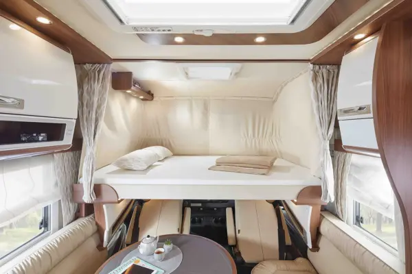 Innenraum eines modernen Carthago Chic E Line Wohnmobils mit einem erhöhten Bett mit Kissen und Decken, einem Essbereich mit Tisch, Teeservice und Zeitschrift, umgeben von beigen Sitzmöbeln und Holzakzenten. Durch die Oberlichter fällt natürliches Licht ein.