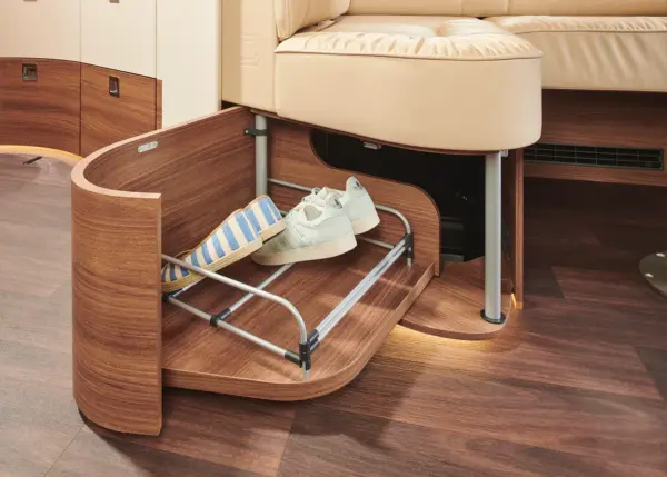Eine ausziehbare Holzschublade unter einem beigen Sofa in einer Carthago Chic E Line enthält weiße Turnschuhe und eine blau gestreifte Tasche mit Metallschienen zur Organisation auf einem dunklen Holzboden.