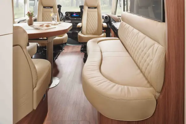 Innenraum eines Carthago Chic E Line Wohnmobils mit hellbraunen Ledersitzen, einem geschwungenen Sofa, einem Esstisch mit Tellern und Gläsern und Bodenbelägen in Holzoptik, die eine gemütliche und moderne Atmosphäre schaffen. Fahrer- und Beifahrersitz sind im Hintergrund zu sehen.