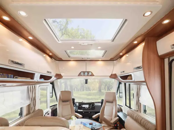 Der Innenraum eines modernen Carthago Chic E Line Wohnmobils ist mit beigen Ledersitzen, Holzakzenten, großen Fenstern mit Vorhängen, Stauraum über dem Kopf und einem Dachfenster ausgestattet, von dem aus man Bäume sehen kann.