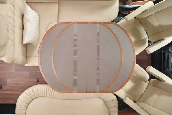 Blick von oben auf den Innenraum eines Carthago Chic E Line Wohnmobils mit einem grauen ovalen Tisch mit Holzakzenten, umgeben von beigen Ledersitzen und einem gemusterten Kissen auf einer Sitzbank. Der Bodenbelag aus dunklem Holz ist teilweise sichtbar.