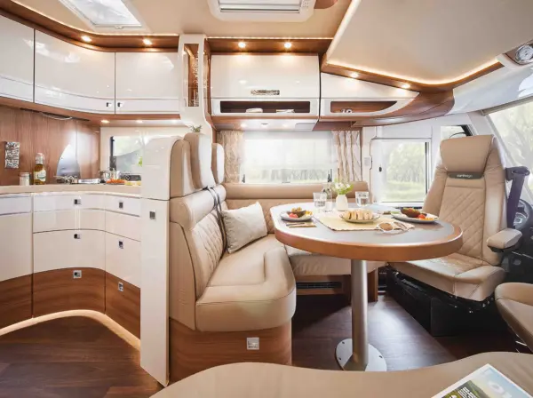 Geräumiges, modernes Wohnmobil-Interieur im Carthago Chic S Plus mit einem gemütlichen Essbereich mit rundem Tisch, beigefarbenen Sitzgelegenheiten, Holzakzenten und einer kompakten Küche mit glänzenden Schränken und natürlichem Licht, das hereinströmt.