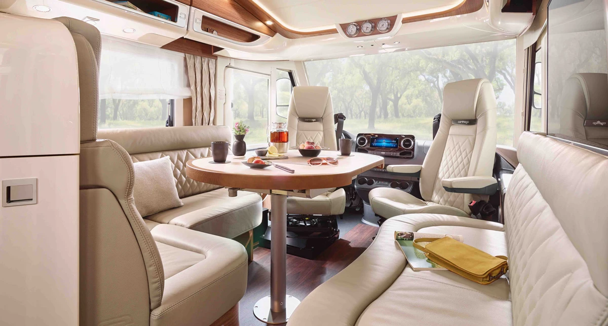 Das moderne Wohnmobil-Interieur im Carthago Chic E Line verfügt über beigefarbene Ledersitze, einen runden Holztisch mit Speisen, große Fenster mit transparenten Vorhängen und Blick auf grüne Bäume. Die Atmosphäre ist hell, gemütlich und einladend.