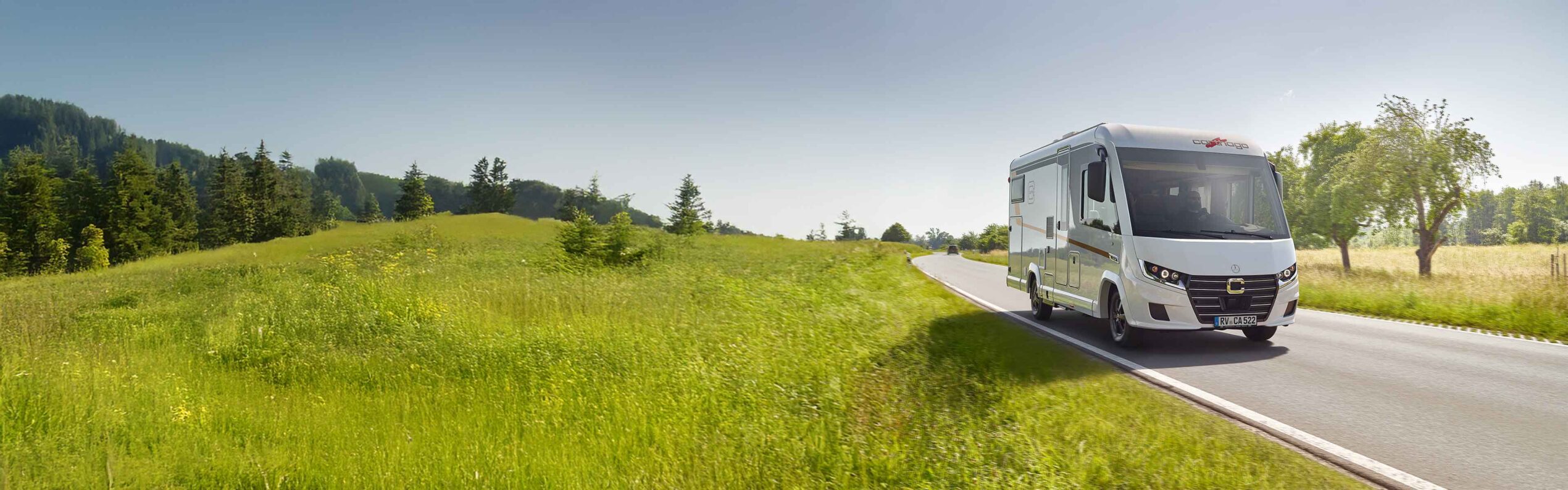 Ein weißes vollintegriertes Wohnmobil fährt auf einer schmalen, kurvenreichen Straße durch eine üppige, grüne Landschaft mit Bäumen und grasbewachsenen Feldern unter einem klaren blauen Himmel.