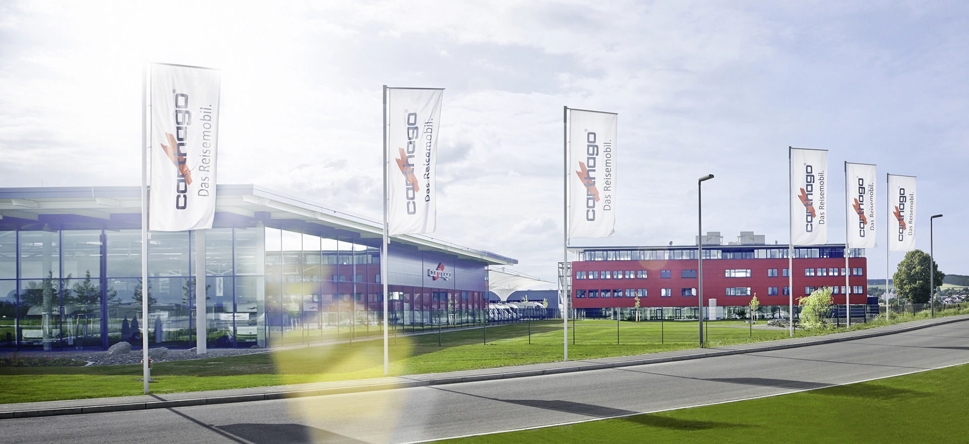 Ein moderner Gebäudekomplex mit großen Fenstern und roten Akzenten steht hinter einer Reihe von sechs hohen weißen Fahnen mit dem Carthago-Logo, neben einer sauberen, leeren Straße unter einem teilweise bewölkten Himmel.