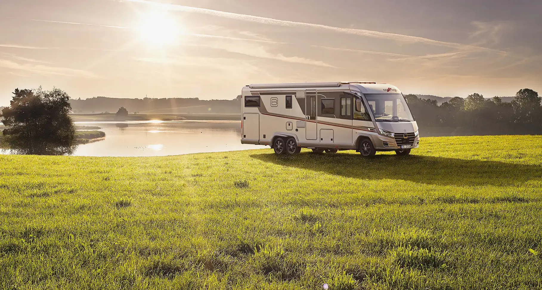 Ein weißes Carthago Wohnmobil ist auf einer grünen Wiese in der Nähe eines ruhigen Sees geparkt, mit Bäumen im Hintergrund. Die Sonne steht tief am Himmel und wirft ein warmes, goldenes Licht über die friedliche Landschaft - der perfekte Ort, um Carthago Wohnmobile Preise zu erkunden.