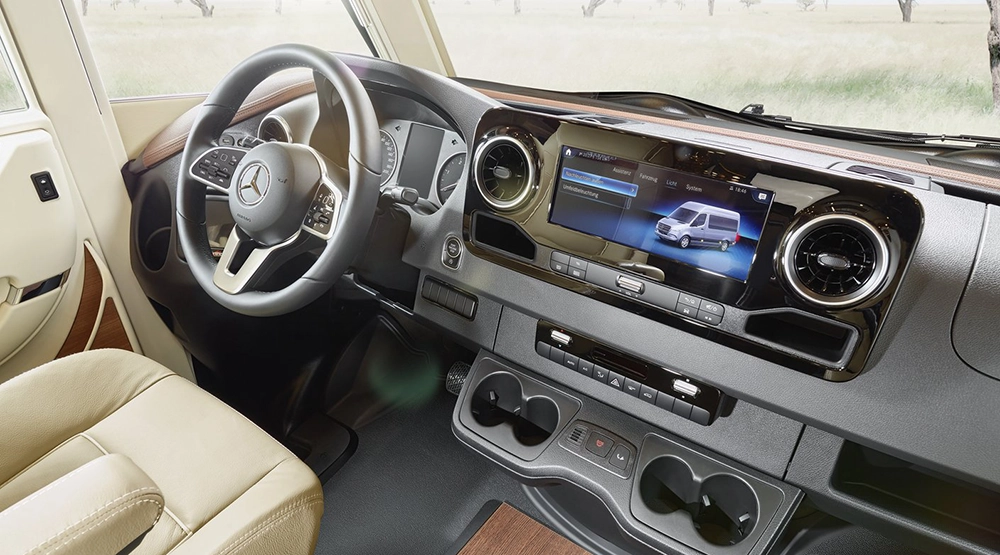 The interior of a Mercedes-Benz vehicle showing the steering wheel, beige leather seats, dashboard and large infotainment screen displaying an image of a van – perfect for a Mercedes motorhome adventure. A grassy meadow is visible though the windows.