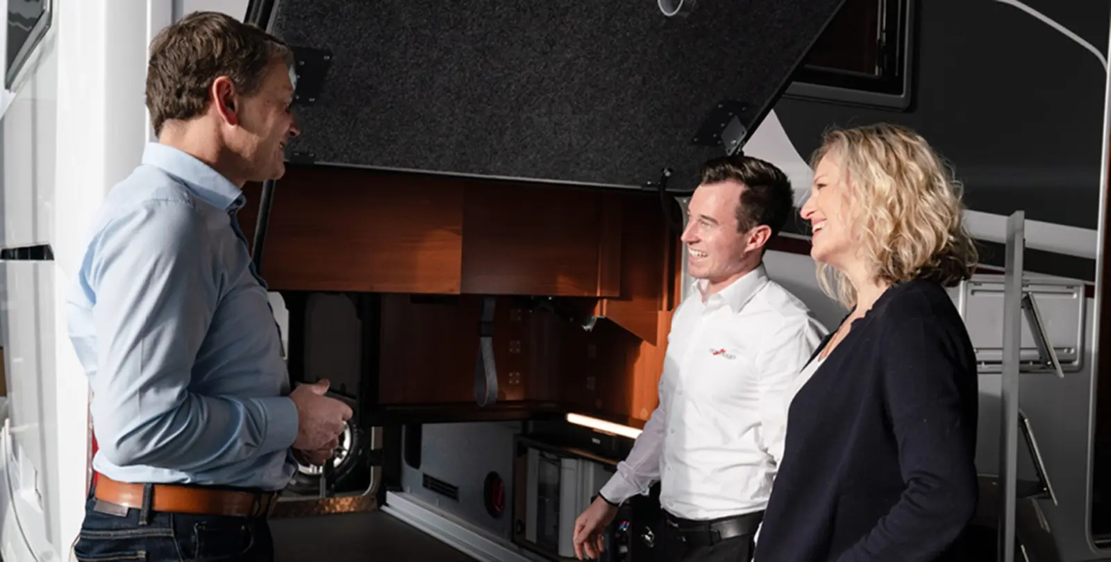 Drei Personen stehen und unterhalten sich in der Nähe eines offenen Stauraums eines Wohnmobils (RV). Zwei von ihnen lächeln, und das Sonnenlicht scheint auf sie und hebt die moderne, elegante Innenausstattung des Wohnmobils im Hintergrund hervor.