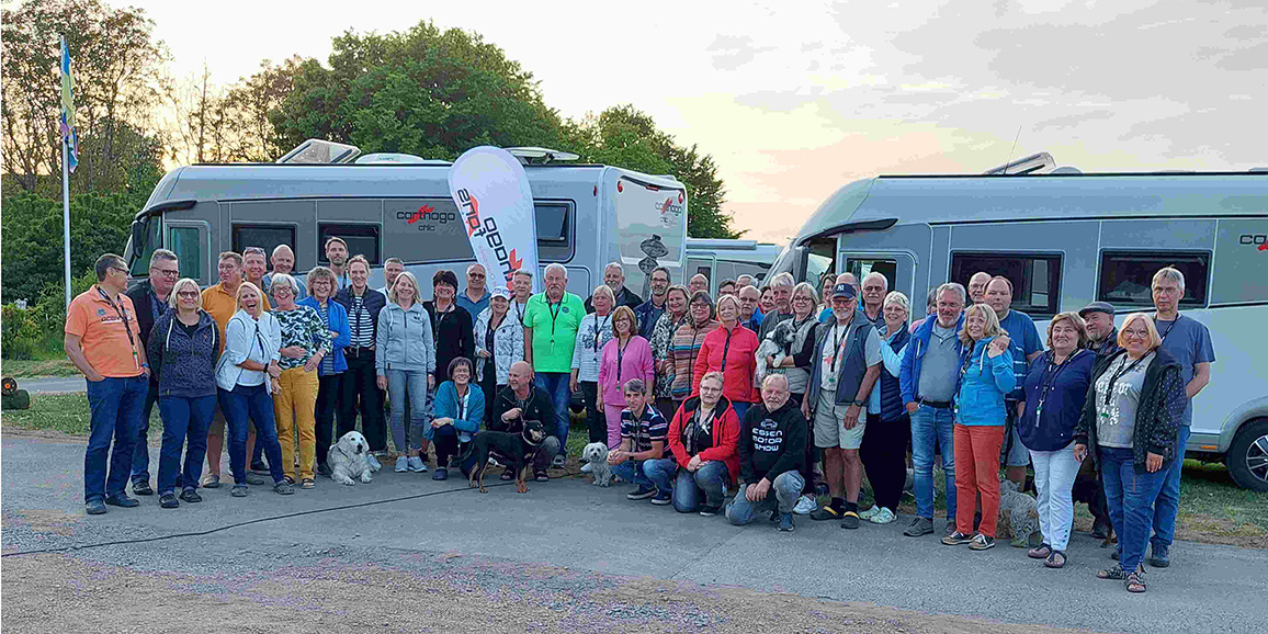 Eine große Gruppe von Menschen unterschiedlichen Alters, die gemeinsam im Freien vor zwei Wohnmobilen posieren, mit Bäumen und einem Sonnenuntergangshimmel im Hintergrund. Einige Leute halten Hunde, und alle lächeln in die Kamera.