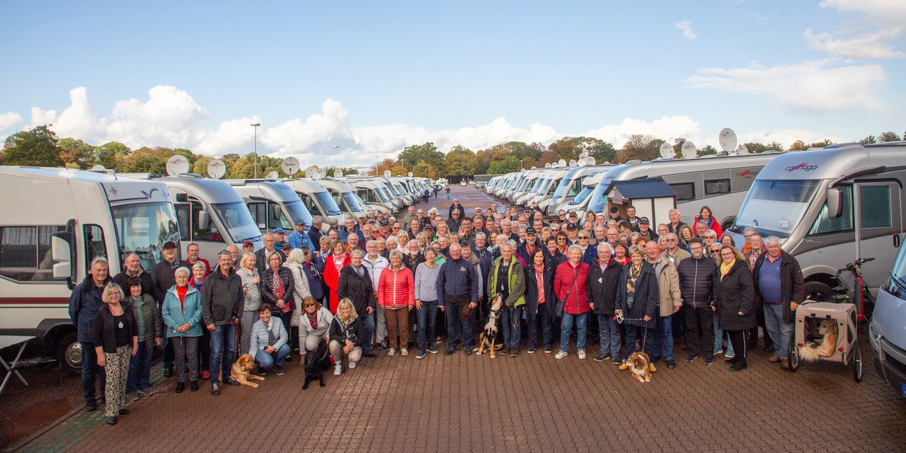 Eine große Gruppe von Menschen und mehrere Hunde stehen lächelnd vor Reihen von geparkten Wohnmobilen auf beiden Seiten einer breiten gepflasterten Fläche, unter einem hellen Himmel mit vereinzelten Wolken.