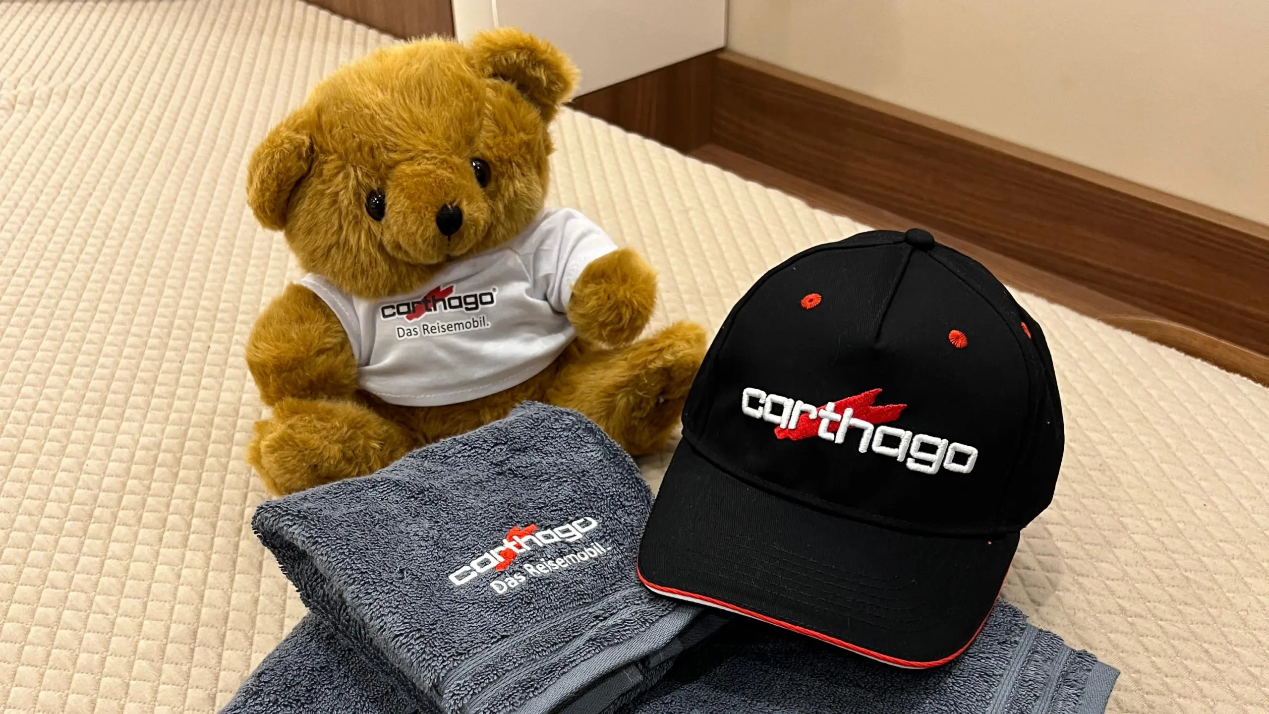 A brown teddy bear wearing a white Carthago shirt sits on a beige carpet next to two grey Carthago towels and a black Carthago baseball cap with red accents.