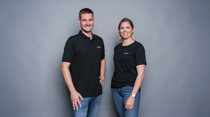 Två personer står bredvid varandra framför en monokrom grå bakgrund, båda ler. De är klädda i svarta T-shirts och blå jeans. Personen till vänster är en man, personen till höger är en kvinna.