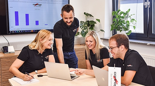 Vier Kollegen in schwarzen Polohemden sitzen und stehen um einen Tisch, lächeln und diskutieren über die Arbeit bei Carthago Slowenien, während sie auf Laptops schauen. Auf einem Bildschirm im Hintergrund ist eine Präsentation mit einem Balkendiagramm zu sehen.