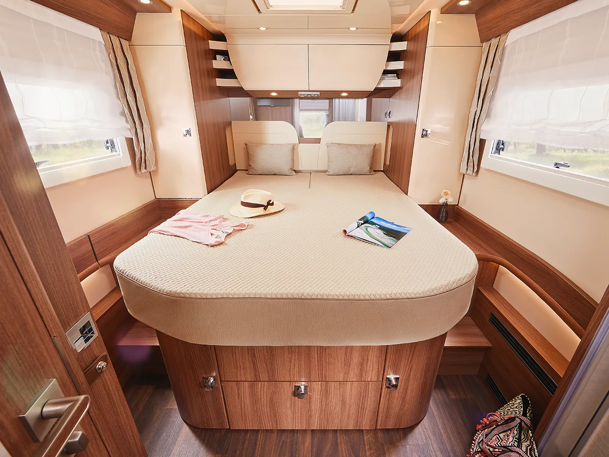 Ein gemütliches Carthago Chic E Line Wohnmobil-Schlafzimmer mit einem großen, runden Bett, Holzschränken, sonnendurchfluteten Fenstern mit Vorhängen, einem Hut, einem Hemd und einer Zeitschrift auf dem Bett sowie einer warmen Deckenbeleuchtung.
