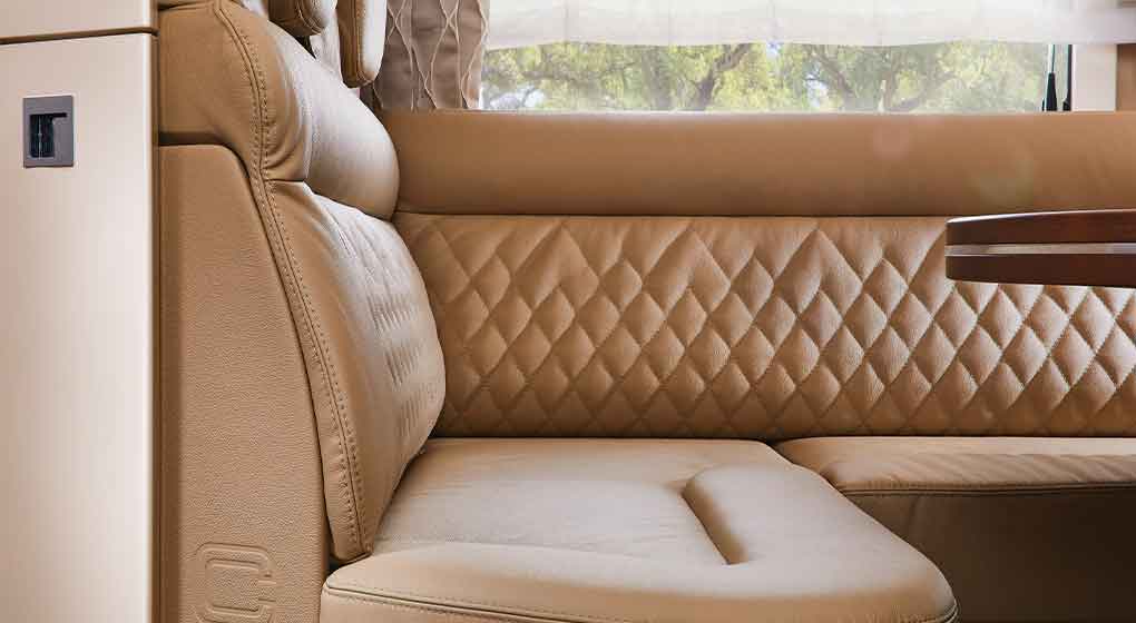 Sièges en cuir marron clair matelassés avec dossier rembourré, dans l’habitacle bien éclairé d’un camping-car ou d’un véhicule ; à l’arrière-plan, on aperçoit une baie avec des rideaux transparents et des plantes vertes à l’extérieur.