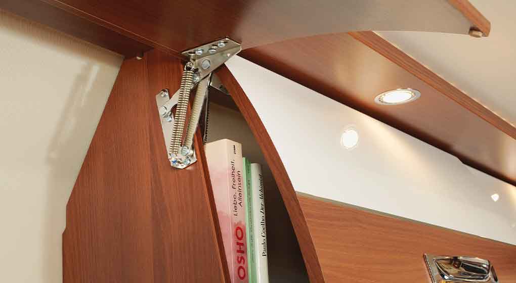 Gros plan sur une porte de placard en bois munie d’une charnière métallique et d’un mécanisme à ressort, partiellement ouverte, laissant apparaître plusieurs livres à l’intérieur. Des lampes encastrées sont installées dans le panneau supérieur au-dessus du placard.