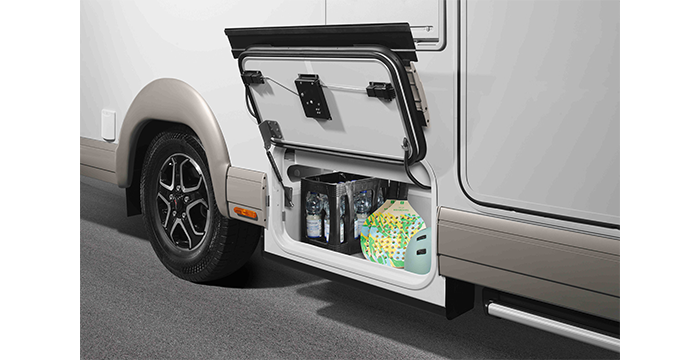 A close-up of the external side compartment of a motorhome that has been opened to reveal items such as bottles of water, a pack of paper towels and a green and white container. The motorhome is parked on a paved area.