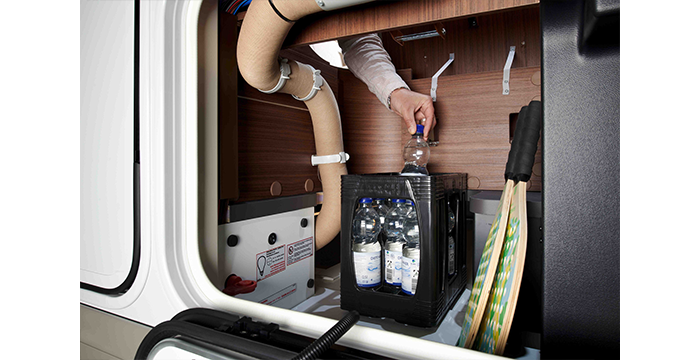 A person filling a water bottle from a dispenser in a compact, well-organised storage compartment containing bottles of water, hoses and cleaning equipment in a vehicle or motorhome.