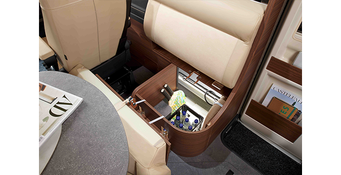 A beige car seat, with its lower section open, reveals a hidden storage compartment containing drinks and snacks. A table with a magazine on top and a door pocket containing books can be seen nearby.