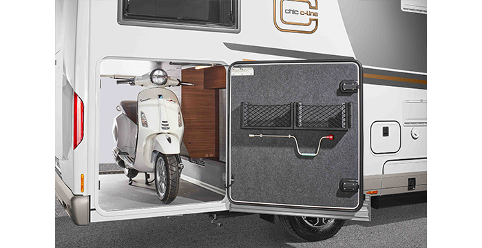 A white scooter is parked in the storage space of a motorhome, with the storage compartment door open to reveal the scooter and a tool holder attached to the inside of the door.