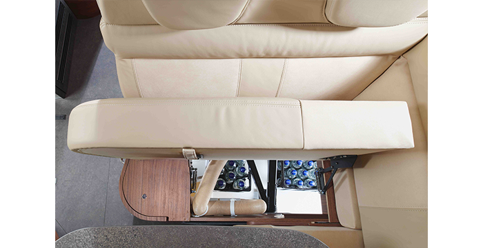 Overhead view of a beige leather sofa in a motorhome, with a storage compartment under the seat containing several bottles of water. Wooden trim and part of a worktop are also visible.