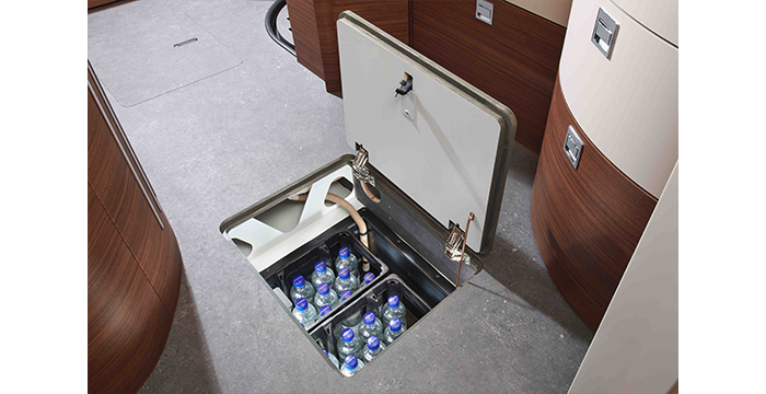 A hidden floor compartment in a modern motorhome reveals neatly arranged bottles of water stored inside. The open hatch is surrounded by wooden trim and grey floor covering.