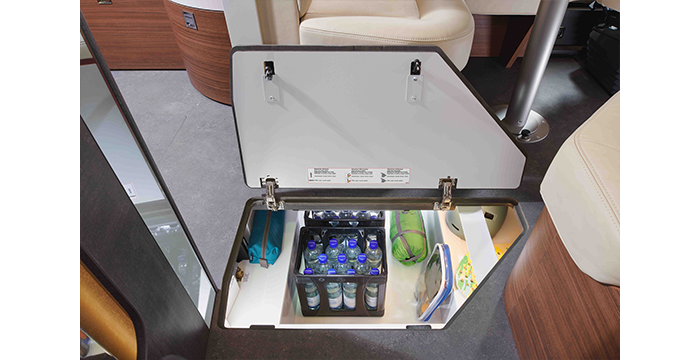 A storage compartment in the floor of a motorhome that can be opened to store bottles of water, drinks and other supplies. The surrounding area is furnished with beige seats and wooden cabinets.