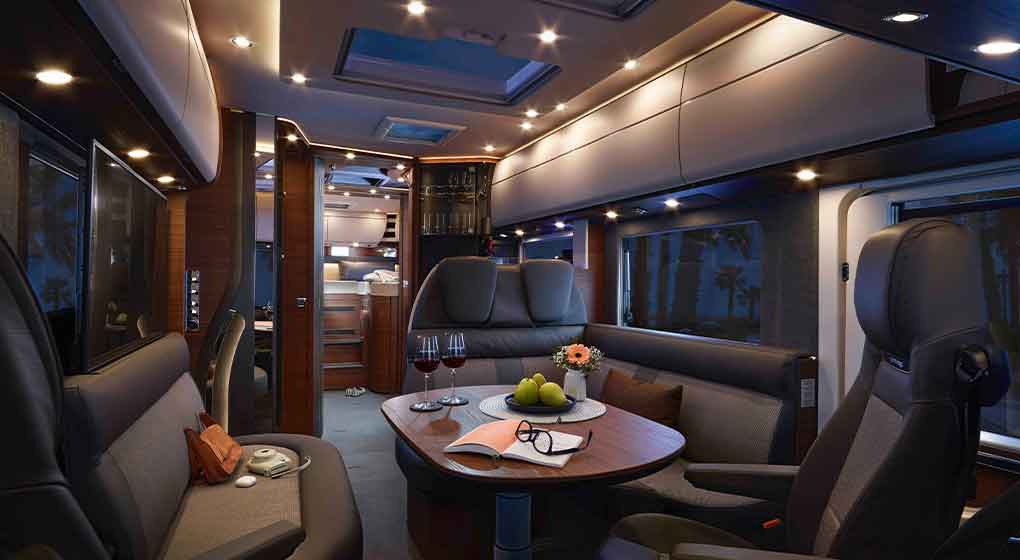 Intérieur luxueux d’un camping-car, avec un éclairage moderne, des sièges en cuir, une table ronde sur laquelle sont disposés des verres à vin, des fruits et un livre. La pièce est dotée de grandes baies, d’espaces de rangement en hauteur et d’une ambiance confortable et accueillante.