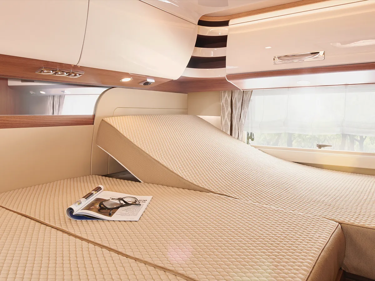 Ein modernes, beigefarbenes verstellbares Bett in einem gut beleuchteten Carthago Chic E Line Wohnmobil mit Holzakzenten. Das Bett ist teilweise erhöht, und eine aufgeschlagene Zeitschrift mit Lesebrille liegt auf der Matratze neben einem Fenster mit durchsichtigen Vorhängen.