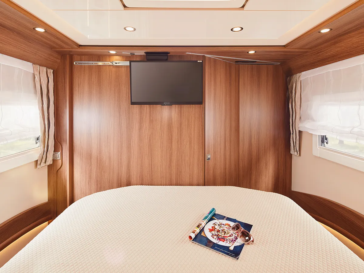 Ein gemütliches Schlafzimmer in der Carthago Chic E Line verfügt über einen an der Wand montierten Fernseher über einem Holzpaneel, ein Bett mit einem strukturierten weißen Bezug und zwei Fenster mit transparenten Vorhängen, die natürliches Licht hereinlassen. Eine Zeitschrift und eine Lesebrille liegen auf dem Bett.